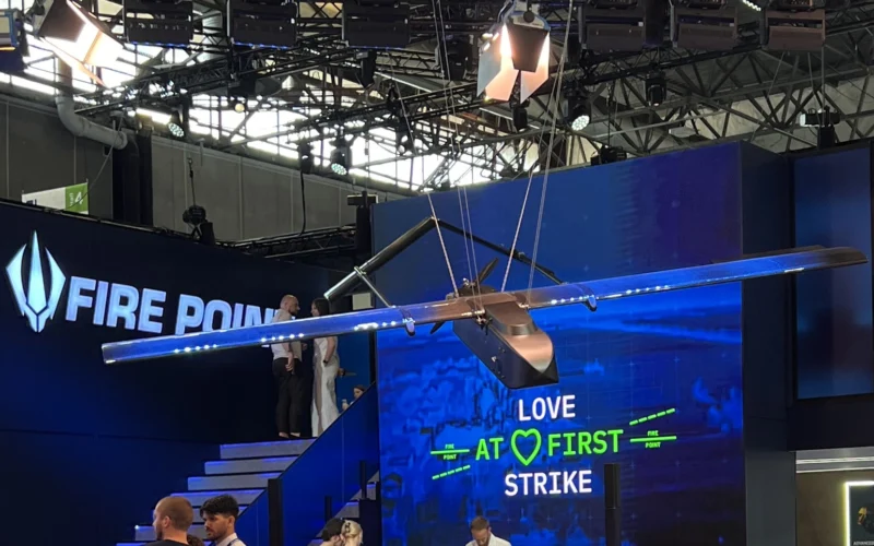 Suspended aircraft wing model overhead at a blue booth with a digital screen reading LOVE AT FIRST STRIKE and FIRE POINT branding