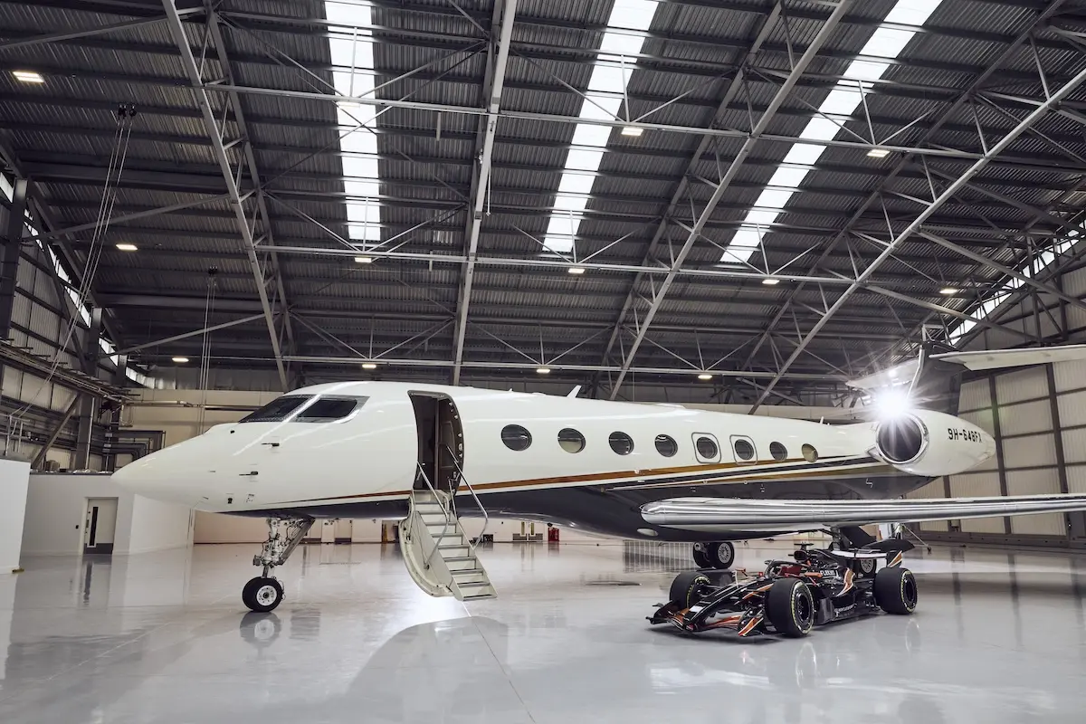 Flexjet and Formula 1® Partnership 2026 White private jet parked inside a spacious hangar with boarding stairs at the open door a race car sits beneath the wing nearby