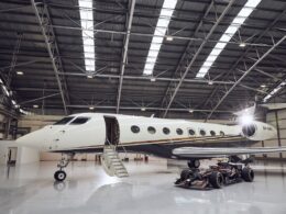 White private jet parked inside a spacious hangar with boarding stairs at the open door a race car sits beneath the wing nearby