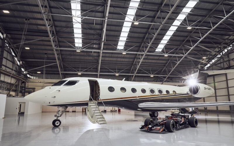 Flexjet and Formula 1® Partnership 2026 White private jet parked inside a spacious hangar with boarding stairs at the open door a race car sits beneath the wing nearby