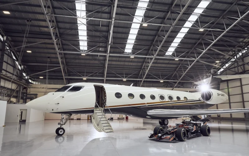 White private jet parked inside a spacious hangar with boarding stairs at the open door a race car sits beneath the wing nearby