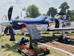 Blue small propeller airplane on display at an outdoor show with open side door and an informational board in front