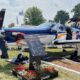 Blue small propeller airplane on display at an outdoor show with open side door and an informational board in front