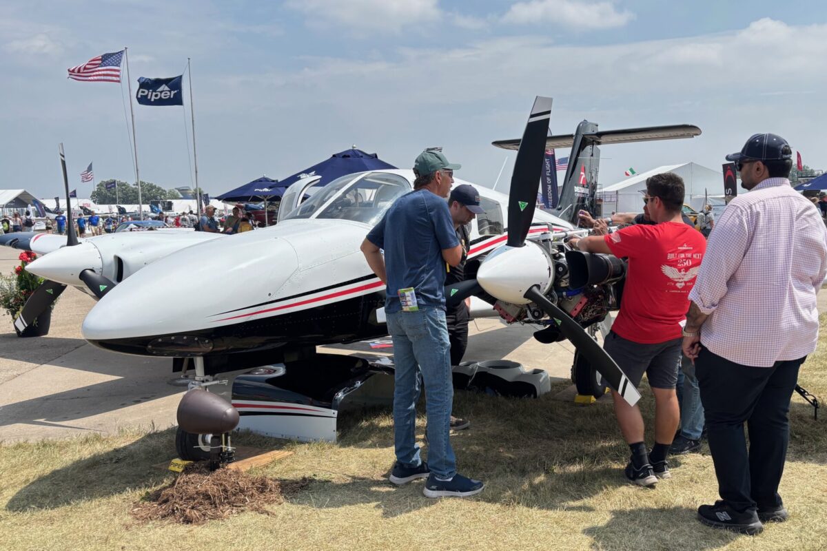 IMG 1282 Group of people inspect a small white propeller aircraft on display at an outdoor airshow with tents and flags in the background under a sunny sky