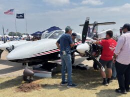 Group of people inspect a small white propeller aircraft on display at an outdoor airshow with tents and flags in the background under a sunny sky