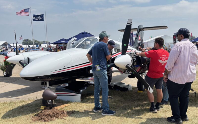 IMG 1282 Group of people inspect a small white propeller aircraft on display at an outdoor airshow with tents and flags in the background under a sunny sky