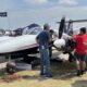 Group of people inspect a small white propeller aircraft on display at an outdoor airshow with tents and flags in the background under a sunny sky