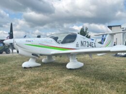 White single engine aircraft with green and red stripes on grass at an airshow tail number N734EX visible on fuselage Elixir
