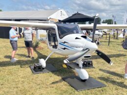 Two men talking beside a white Pipistrel aircraft on display at an outdoor aviation expo with tents and attendees in the background