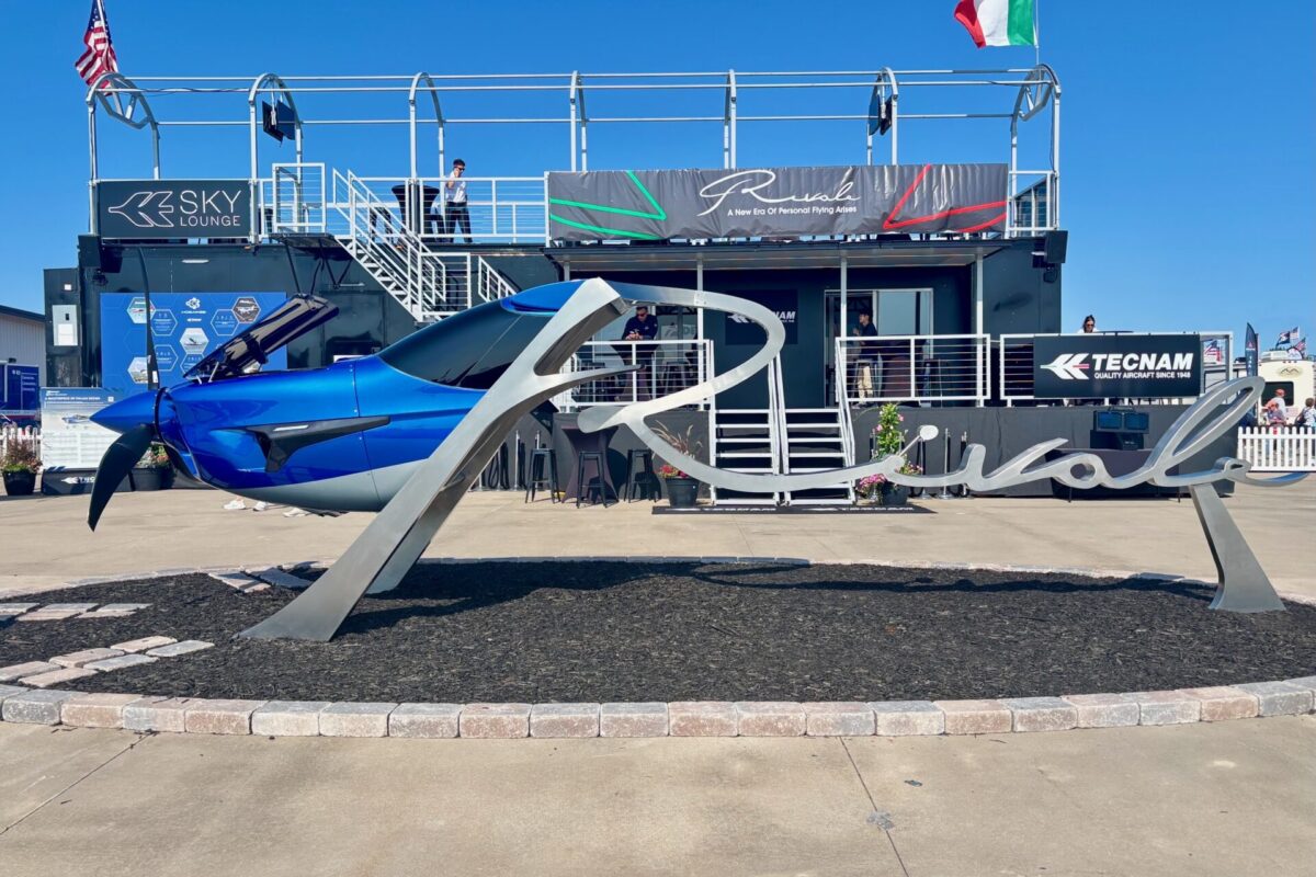 Blue aircraft on a display pedestal at an outdoor aviation show with a two story exhibition structure and flags in the background