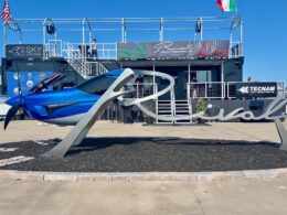 Blue aircraft on a display pedestal at an outdoor aviation show with a two story exhibition structure and flags in the background