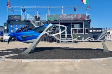 Blue aircraft on a display pedestal at an outdoor aviation show with a two story exhibition structure and flags in the background