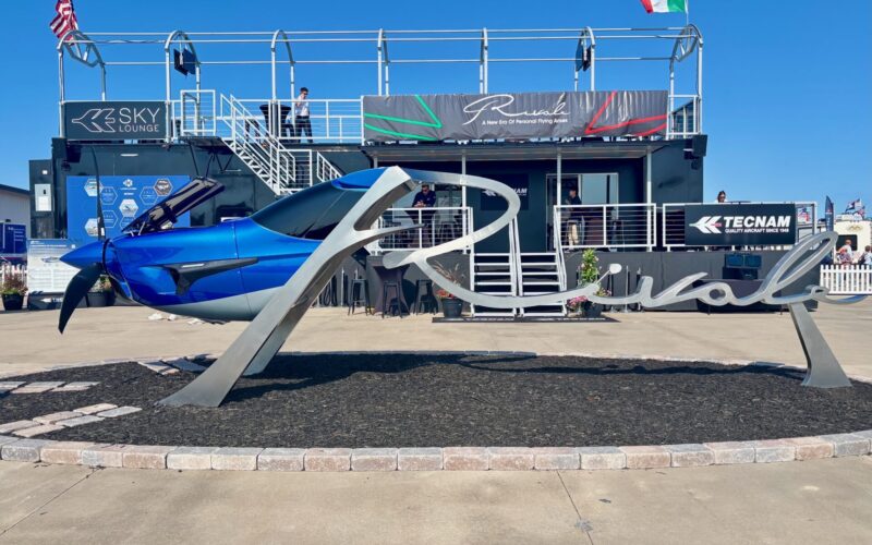 IMG 1318 Blue aircraft on a display pedestal at an outdoor aviation show with a two story exhibition structure and flags in the background