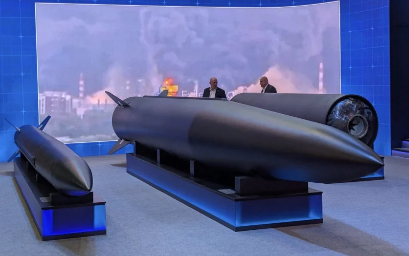 Exhibition display a large dark missile on a stand with two suited figures discussing it blue backdrop reading 18062026 MOSCOW OIL REFINERY