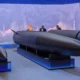 Exhibition display a large dark missile on a stand with two suited figures discussing it blue backdrop reading 18062026 MOSCOW OIL REFINERY