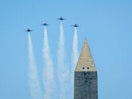 Four jets fly in tight formation over a tall obelisk leaving white smoke trails against a clear blue sky