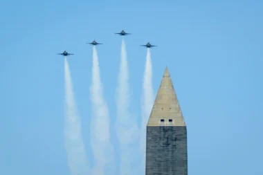 Four jets fly in tight formation over a tall obelisk leaving white smoke trails against a clear blue sky