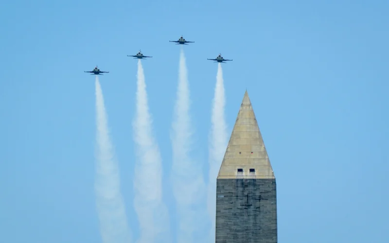 Four jets fly in tight formation over a tall obelisk leaving white smoke trails against a clear blue sky