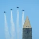 Four jets fly in tight formation over a tall obelisk leaving white smoke trails against a clear blue sky