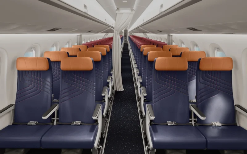 Inside a commercial airplane cabin with two columns of blue seats and orange headrests along a narrow aisle viewed from the rear toward the front carpeted floor visible beneath the seats