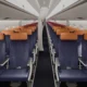 Inside a commercial airplane cabin with two columns of blue seats and orange headrests along a narrow aisle viewed from the rear toward the front carpeted floor visible beneath the seats