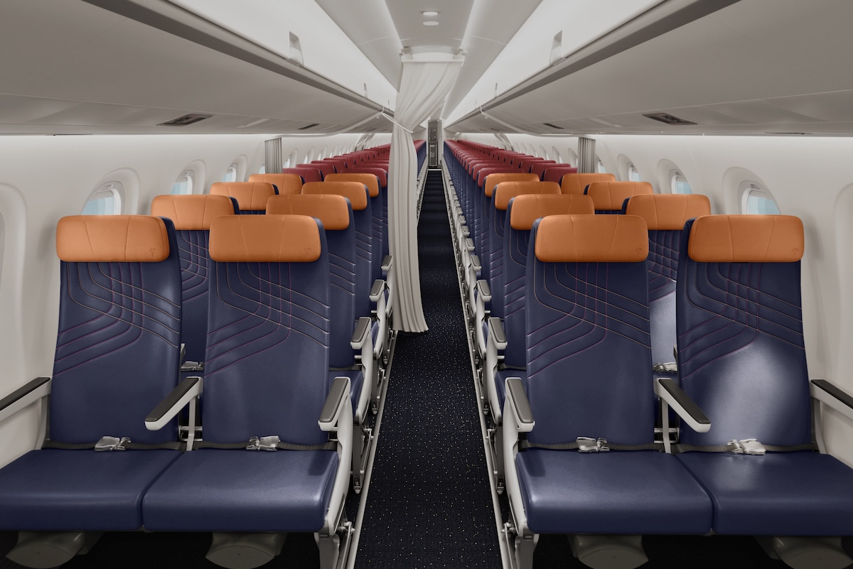 Prem Economy with OPEN MCD show transition Inside a commercial airplane cabin with two columns of blue seats and orange headrests along a narrow aisle viewed from the rear toward the front carpeted floor visible beneath the seats