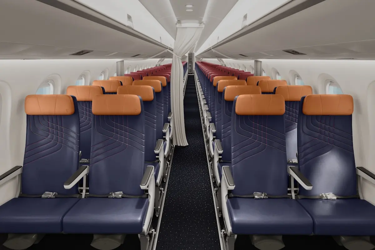 Inside a commercial airplane cabin with two columns of blue seats and orange headrests along a narrow aisle viewed from the rear toward the front carpeted floor visible beneath the seats