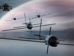 Formation of several drone aircraft with cross shaped wing extensions flying in a hazy sky a helicopter visible in the upper left corner