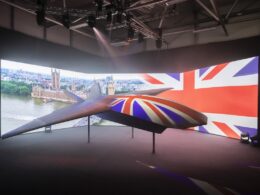 Suspended futuristic aircraft on display indoors with a large Union Jack backdrop and cityscape projection behind it