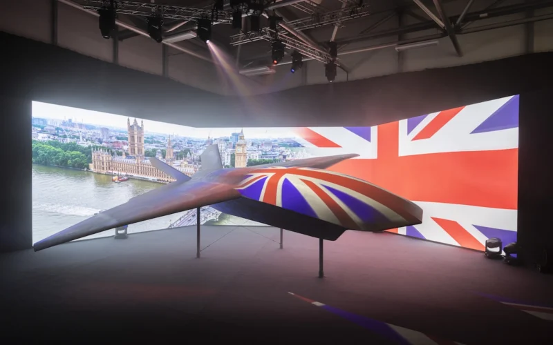 Suspended futuristic aircraft on display indoors with a large Union Jack backdrop and cityscape projection behind it