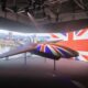 Suspended futuristic aircraft on display indoors with a large Union Jack backdrop and cityscape projection behind it