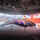 Suspended futuristic aircraft on display indoors with a large Union Jack backdrop and cityscape projection behind it