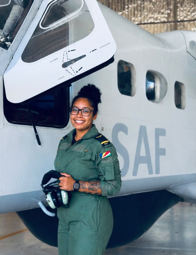 Sylvie Tamboo Female pilot in a green flight suit stands beside a white aircraft holding a helmet and smiling at the camera