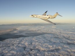 White Gulfstream G800 private jet flying above a sea of clouds with a distant mountain range and a pale sunrise sky