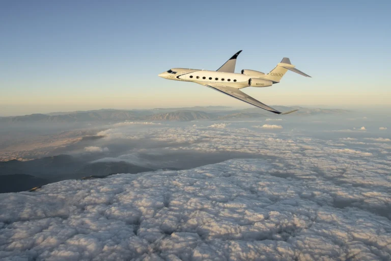 White Gulfstream G800 private jet flying above a sea of clouds with a distant mountain range and a pale sunrise sky