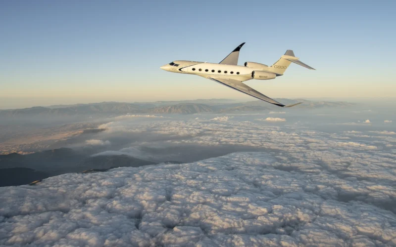 White Gulfstream G800 private jet flying above a sea of clouds with a distant mountain range and a pale sunrise sky