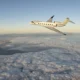 White Gulfstream G800 private jet flying above a sea of clouds with a distant mountain range and a pale sunrise sky