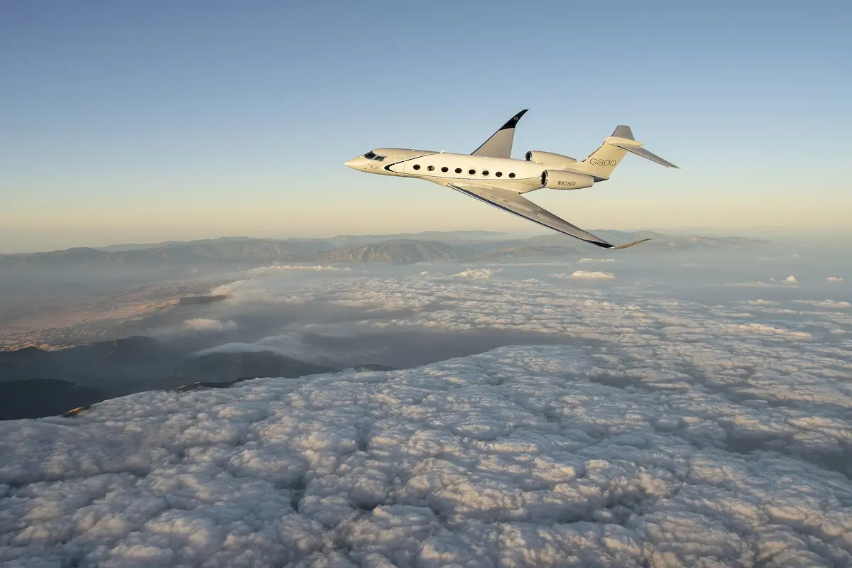 Gulfstream G800 White Gulfstream G800 private jet flying above a sea of clouds with a distant mountain range and a pale sunrise sky