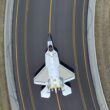 Top down view of a white fighter jet on a dark runway with yellow centerlines surrounded by grassy embankments on both sides