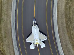 Top down view of a white fighter jet on a dark runway with yellow centerlines surrounded by grassy embankments on both sides