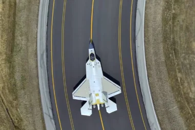 Top down view of a white fighter jet on a dark runway with yellow centerlines surrounded by grassy embankments on both sides