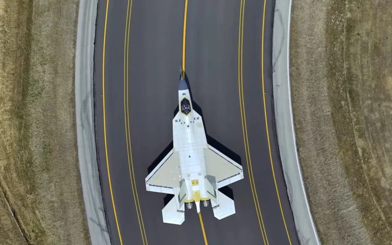 Top down view of a white fighter jet on a dark runway with yellow centerlines surrounded by grassy embankments on both sides