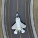 Top down view of a white fighter jet on a dark runway with yellow centerlines surrounded by grassy embankments on both sides