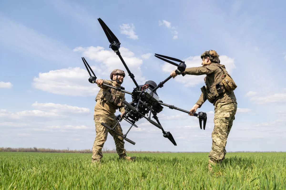 Two soldiers in camouflage lift a large quadcopter drone between them in a grassy field under a blue sky