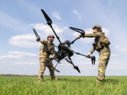 Two soldiers in camouflage lift a large quadcopter drone between them in a grassy field under a blue sky