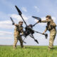 Two soldiers in camouflage lift a large quadcopter drone between them in a grassy field under a blue sky