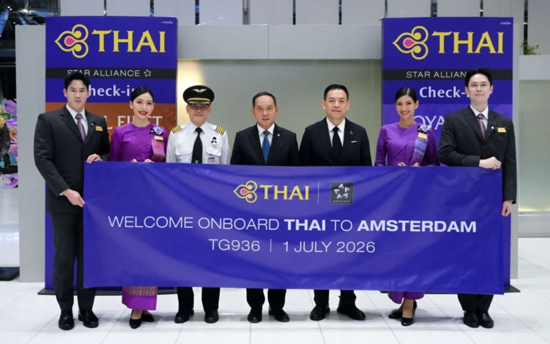 Group of six individuals in formal attire posing behind a large THAI banner at a THAI event Amsterdam welcome sign visible