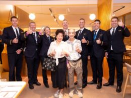 Group of staff in navy uniforms with gold stripes pose with two smiling elderly guests all giving thumbs up in a warmly lit restaurant setting