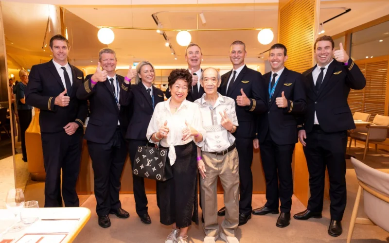 Group of staff in navy uniforms with gold stripes pose with two smiling elderly guests all giving thumbs up in a warmly lit restaurant setting