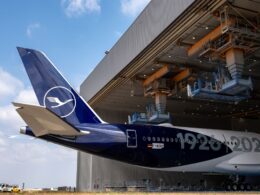 Lufthansa aircraft tail with logo near a large maintenance hangar and overhead equipment
